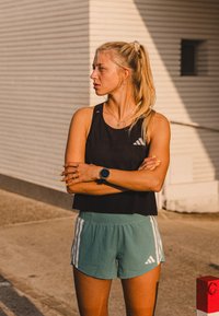 Zwarte sport tanktop met een mesh-textuur; lichtgroene Shorts met witte strepen; smartwatch om de pols; blond haar in een paardenstaart.