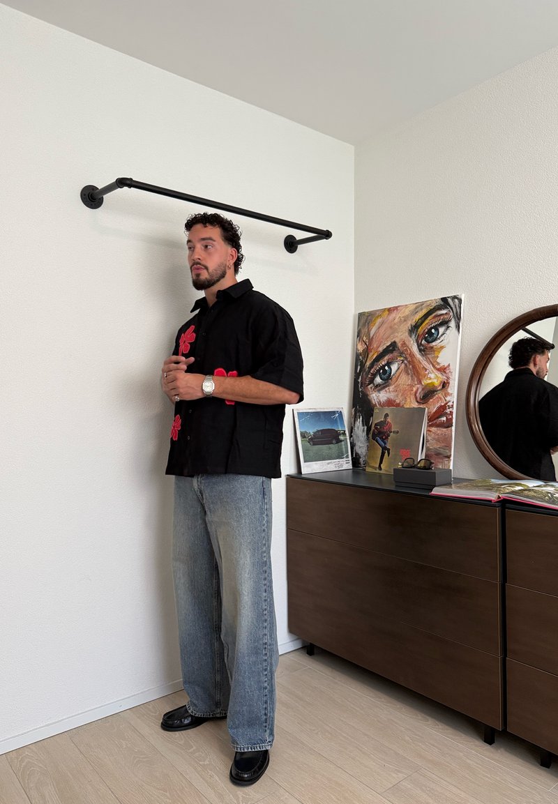 Homme aux cheveux bouclés portant une chemise noire à fleurs rouges et un jean, se tient à côté d'une commode en bois avec des œuvres d'art et un miroir rond.