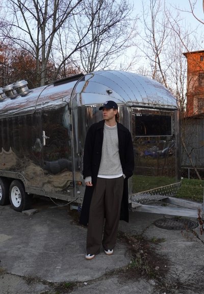 Remolque de aluminio con superficie brillante y techo redondeado. La figura lleva un abrigo negro, suéter gris, pantalones marrones y una gorra oscura, de pie de manera casual.