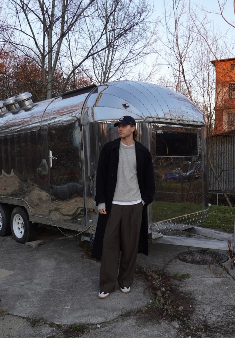 Rimorchio in alluminio con superficie lucida e tetto arrotondato. La figura indossa un cappotto nero, un maglione grigio, pantaloni marroni e un berretto scuro, in posizione rilassata.