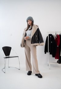 Jaqueta de couro forrada com pele de carneiro preta, calças beje claro de cintura alta, gorro de malha cinza, sapatos plataforma pretos, tudo em um cenário minimalista.