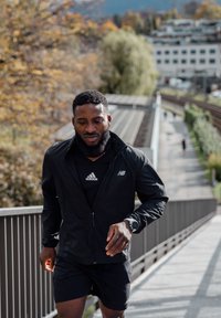 Man wearing a black athletic jacket and shorts jogging along a sunny outdoor path, with railings and autumn trees in the background.