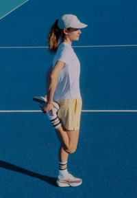 Sporta apavi, maigi gaiši pelēki ar purpura akcentiem; dzelteni šorti; balts T-krekls; svītrainas zeķes. Stiepjas zilajā tenisa kortā.