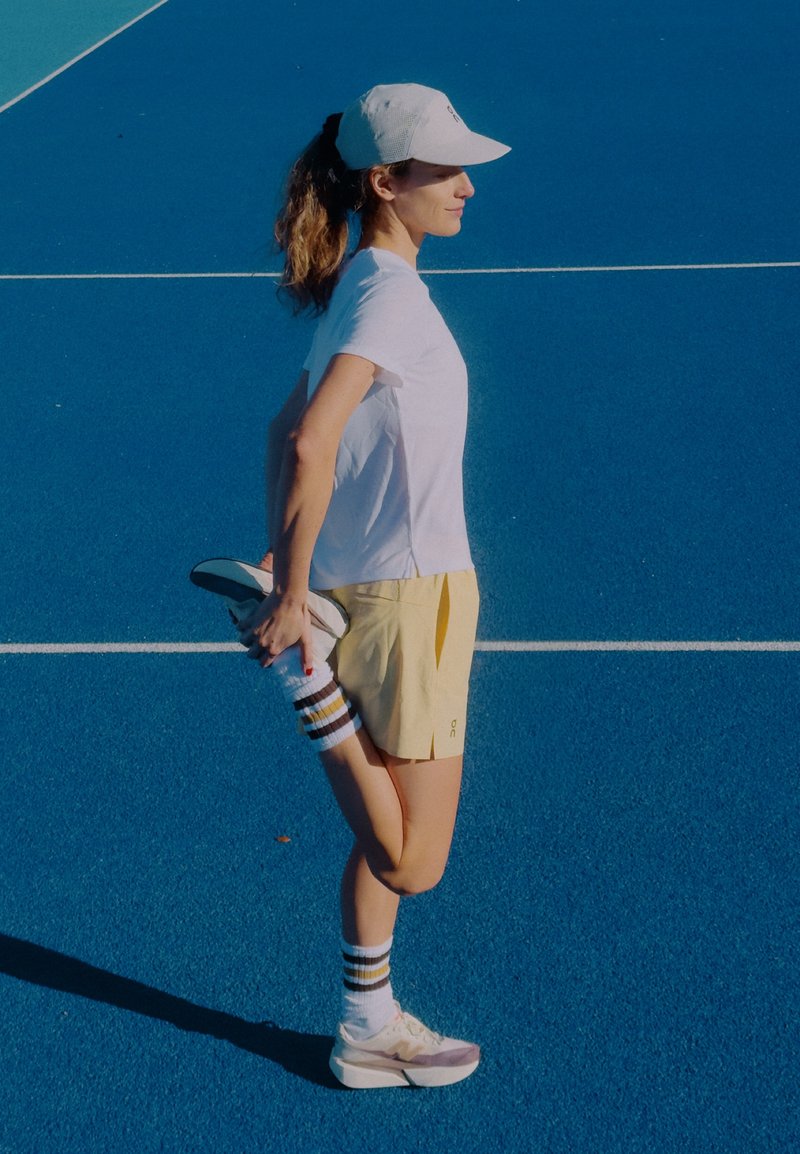 Urheilukengät, pehmeän vaaleanharmaat violetein yksityiskohdin; keltaiset shortsit; valkoinen T-paita; raidalliset sukat. Venytellä sinisellä tenniskentällä.