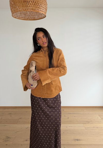 Mujer con cabello largo y oscuro sosteniendo un bolso beige, vestida con una camisa de pana marrón y una falda marrón oscuro con lunares, de pie sobre un suelo de madera bajo una pantalla de lámpara tejida.