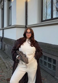 Beige strikket antrekk med rund hals; lagvis kombinert med en mørk brun trenchcoat. Holder en grå teksturert veske, stående mot en lys vegg.