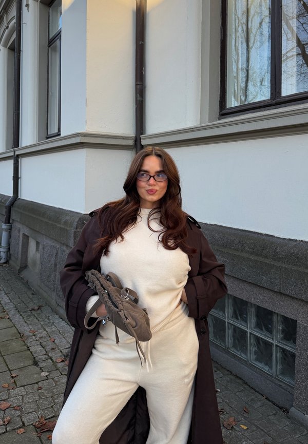Beiges Strickoutfit mit Rundhalsausschnitt; darüber ein dunkelbrauner Trenchcoat. Eine graue, strukturierte Tasche haltend, vor einer hellen Wand stehend.