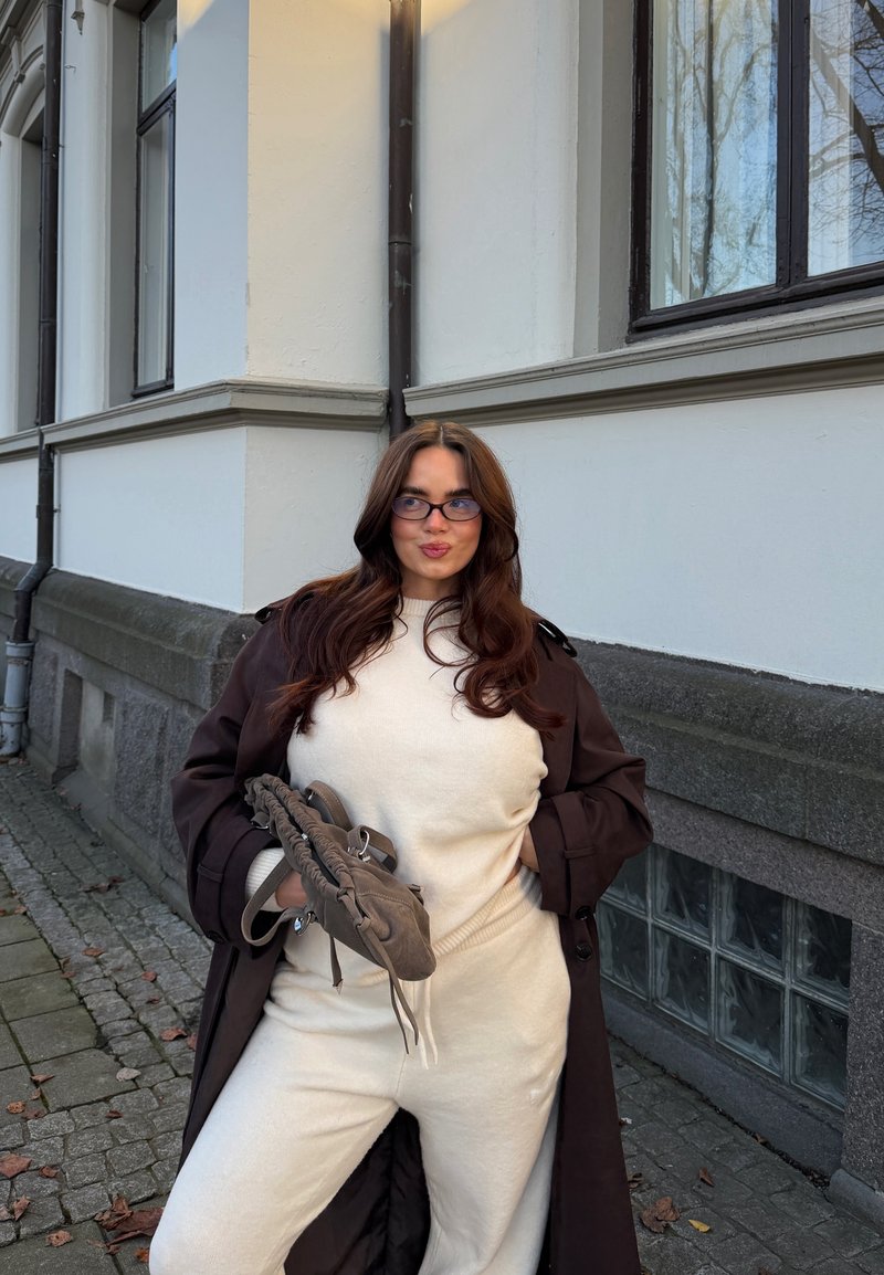Beiges Strickoutfit mit Rundhalsausschnitt; geschichtet mit einem dunkelbraunen Trenchcoat. Eine graue, strukturierte Tasche haltend, steht vor einer hellen Wand.