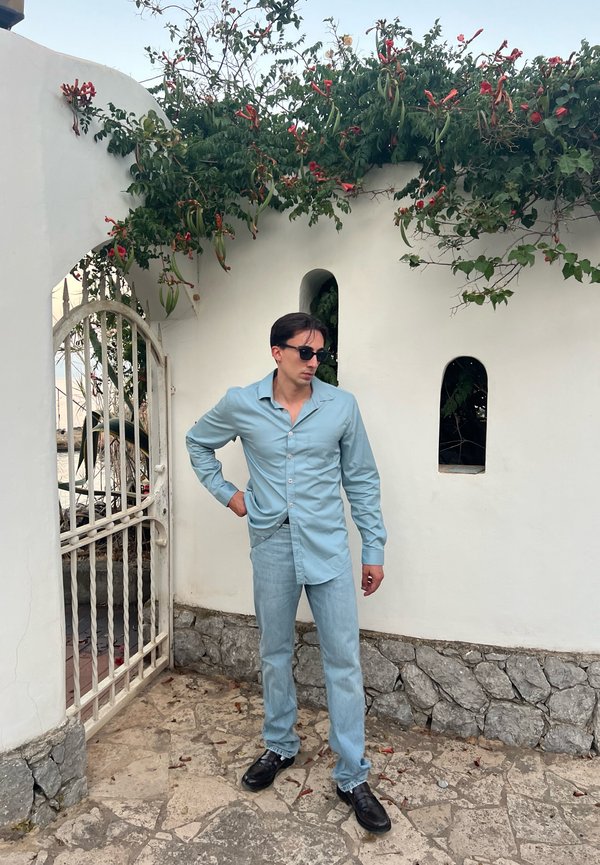 Lichtblauw overhemd met lange mouwen en een kraag, gecombineerd met lichte jeans en zwarte schoenen; geplaatst voor een witte muur met groenigheid.