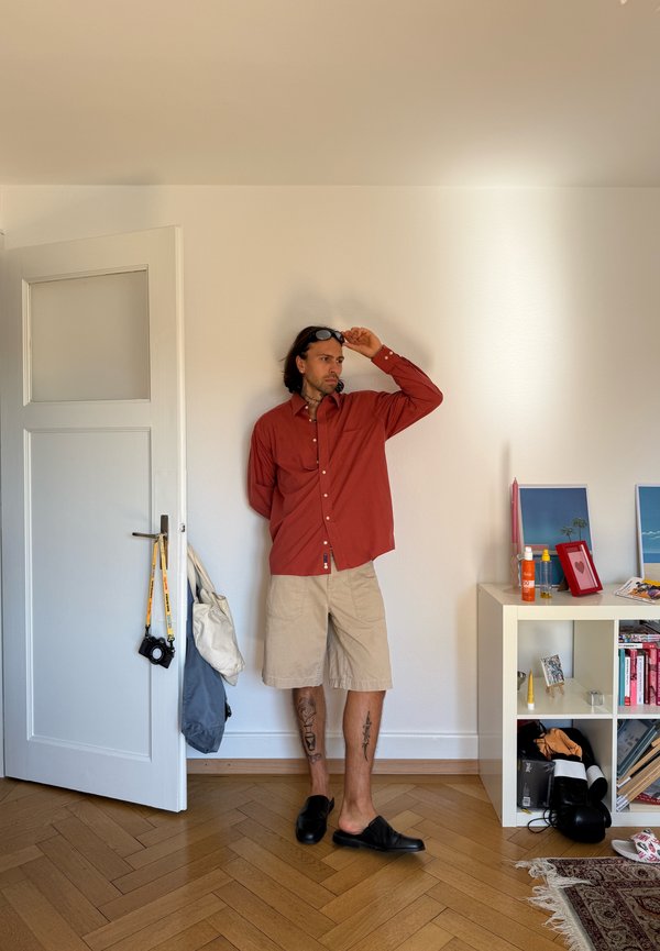 Homme portant une chemise rouge et un short beige, debout contre un mur blanc à l'intérieur, ajustant ses lunettes de soleil d'une main, près d'une porte partiellement ouverte et d'une étagère.
