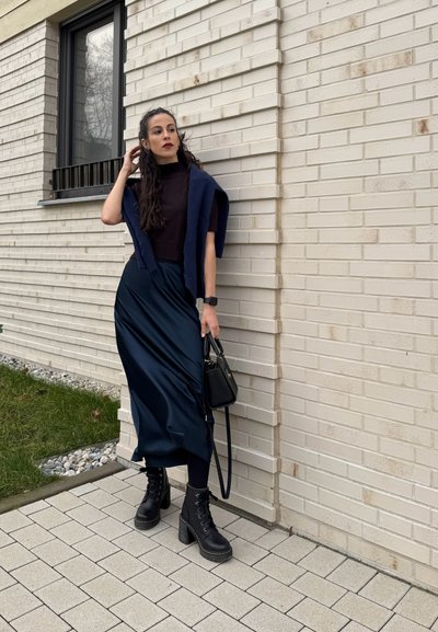 Botas de tacón alto negras con cordones, falda maxi de satén azul marino, blusa ajustada oscura y un chal de punto azul marino. Bolsa negra y fondo de pared de ladrillo texturizado.