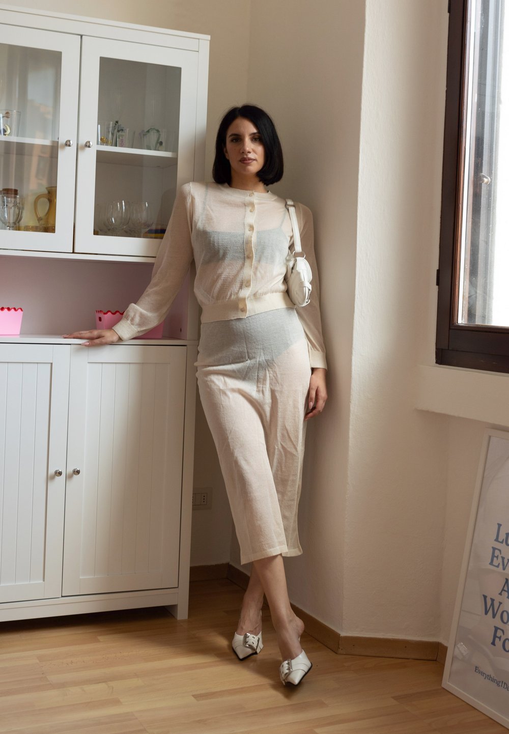 Ανοιχτό μπεζ διπλό σύνολο με διαφανή μακρυμάνικη μπλούζα και εφαρμοστά παντελόνια. Λευκά παπούτσια με τετραγωνική μύτη και φιόγκο. Μινιμαλιστικός χώρος.