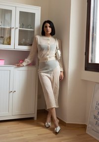 Ανοιχτό μπεζ διπλό σύνολο με διαφανή μακρυμάνικη μπλούζα και εφαρμοστά παντελόνια. Λευκά παπούτσια με τετραγωνική μύτη και φιόγκο. Μινιμαλιστικός χώρος.