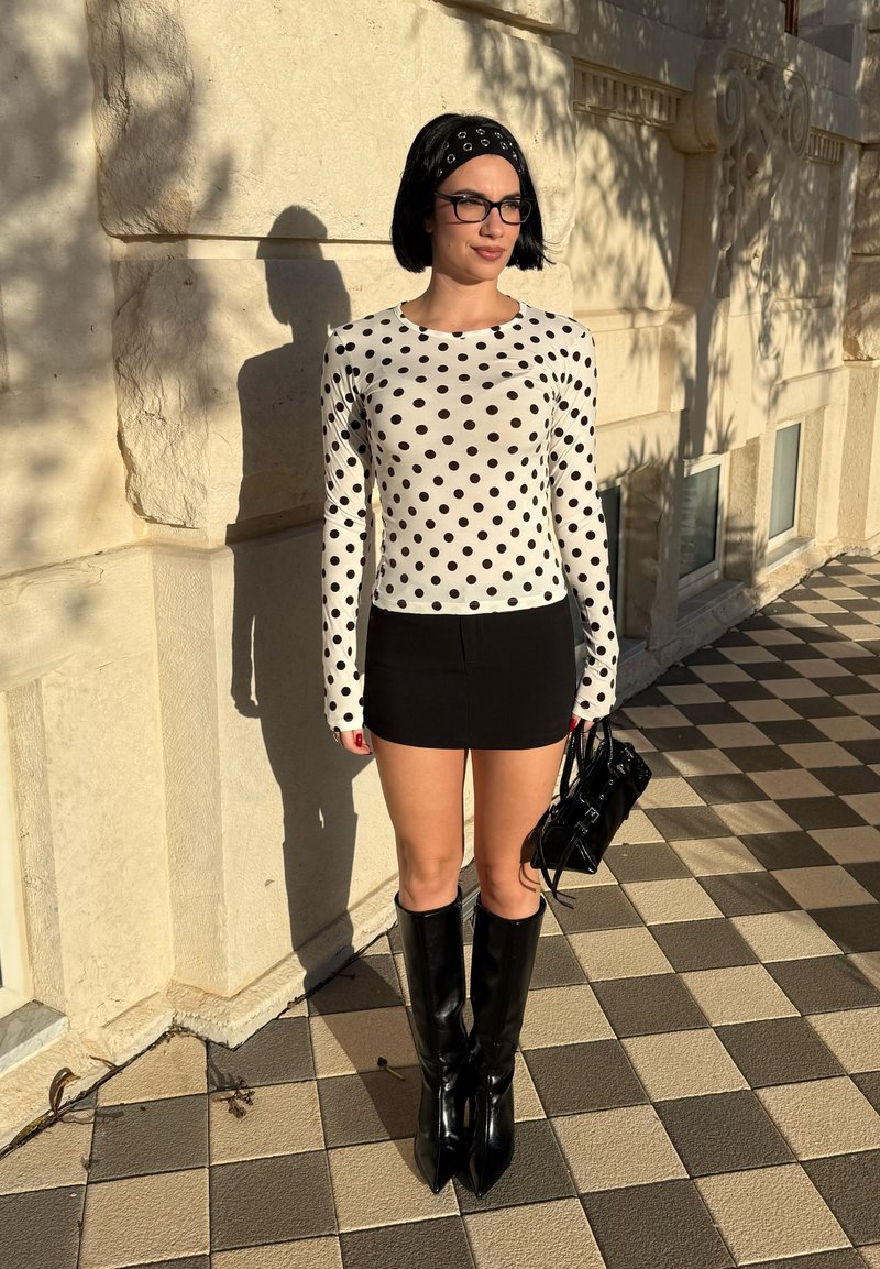 Femme avec un carré noir et des lunettes, portant un haut blanc à manches longues à pois, une jupe noire, des bottes hautes jusqu'aux genoux, et tenant un sac à main noir en extérieur.