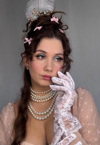 Una donna indossa una collana di perle, guanti di pizzo bianchi e un vestito floreale rosa con maniche trasparenti. Ha un'accessorio per capelli piumato e i capelli acconciati.