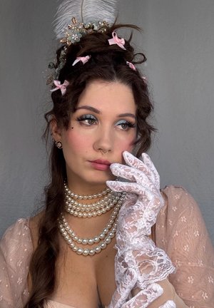 Una mujer lleva un collar de perlas, guantes de encaje blanco y un vestido rosa con flores y mangas transparentes. Tiene una diadema de plumas y el cabello peinado.