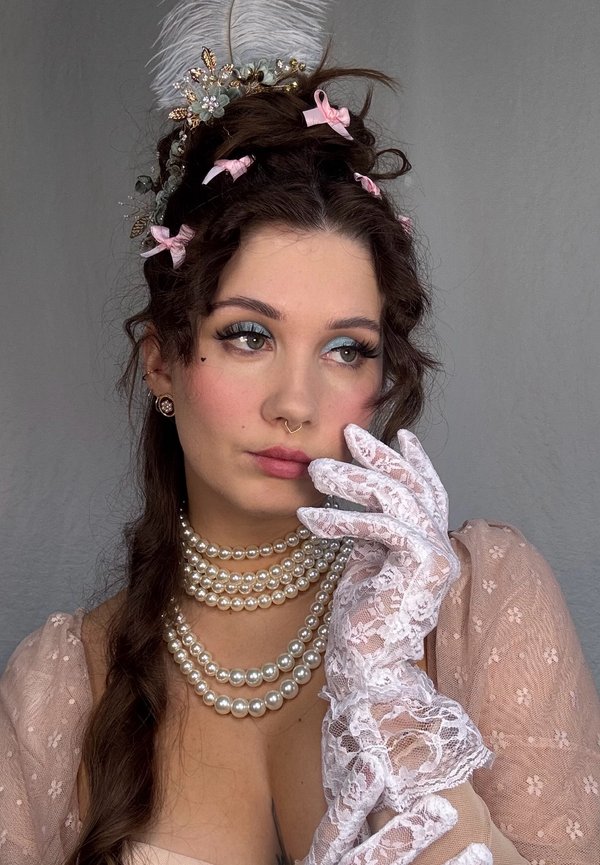 Eine Frau trägt eine Perlenkette, weiße Spitzenhandschuhe und ein pinkfarbenes Blumenkleid mit durchsichtigen Ärmeln. Sie hat ein gefiedertes Haarschmuckstück und gestylte Haare.