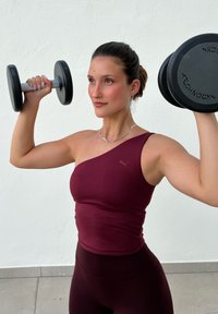 Kvinna i vinröd träningsoutfit som lyfter svarta hantlar inomhus mot en vit vägg.