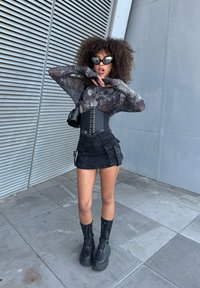 Maglietta nera in rete a maniche lunghe con un motivo scintillante, corsetto nero con lacci, mini gonna nera con tasche e stivali neri chunky con lacci.