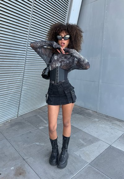 Top de malla negra de manga larga con un patrón brillante, corsé negro con cordones, minifalda negra con bolsillos y botas negras de cordones gruesos.