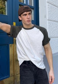 Weißes T-Shirt mit dunkelgrünen Raglanärmeln, kombiniert mit schwarzen Jeans. Im Hintergrund befindet sich eine blaue Tür mit einem vergoldeten Türgriff.