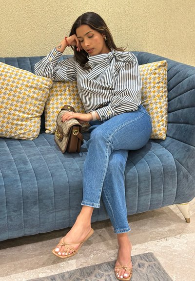 Blusa de rayas con detalle de lazo, vaqueros azules, sandalias beige. Sentada en un sofá azul acolchado con cojines en pata de gallo amarillos y blancos.