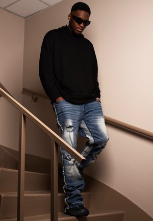 Sudadera negra con un bolsillo delantero, jeans azules desgastados con bordes deshilachados y zapatos negros. De pie en una escalera con paredes beige.