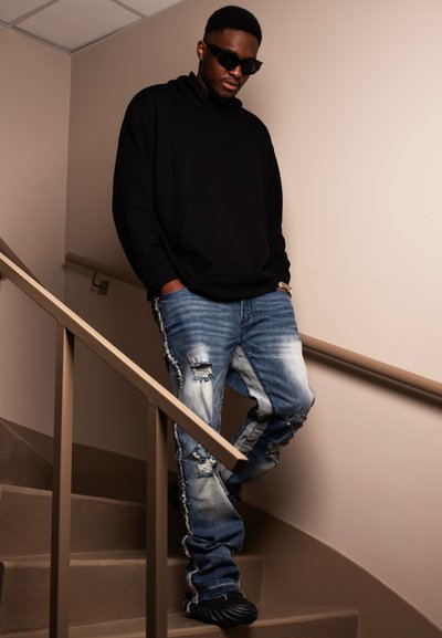 Sudadera negra con un bolsillo delantero, jeans azules desgastados con bordes deshilachados y zapatos negros. De pie en una escalera con paredes beige.