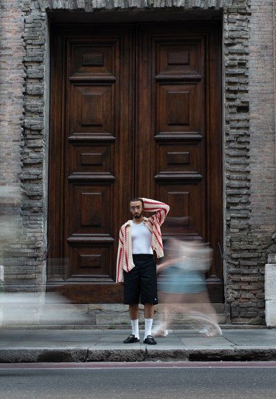 Cárdigan de punto con rayas rojas, camiseta blanca sin mangas, pantalones cortos negros y zapatos negros. El fondo presenta grandes puertas de madera y una pared de piedra.