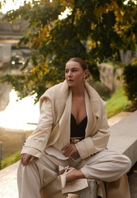 Beige ylbig blazer með mjúku sjalkraga, svört topp, breiðar buxur og beige hælaskór með bogum. Náttúruleg umgjörð, hlý ljós.