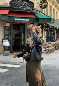 Musta nahkatakki, oliivinvihreä hame sivuhalkiolla ja pieni ruskea käsilaukku. Kahvilan taustalla vihreä markiisi ja ulkoterassi.