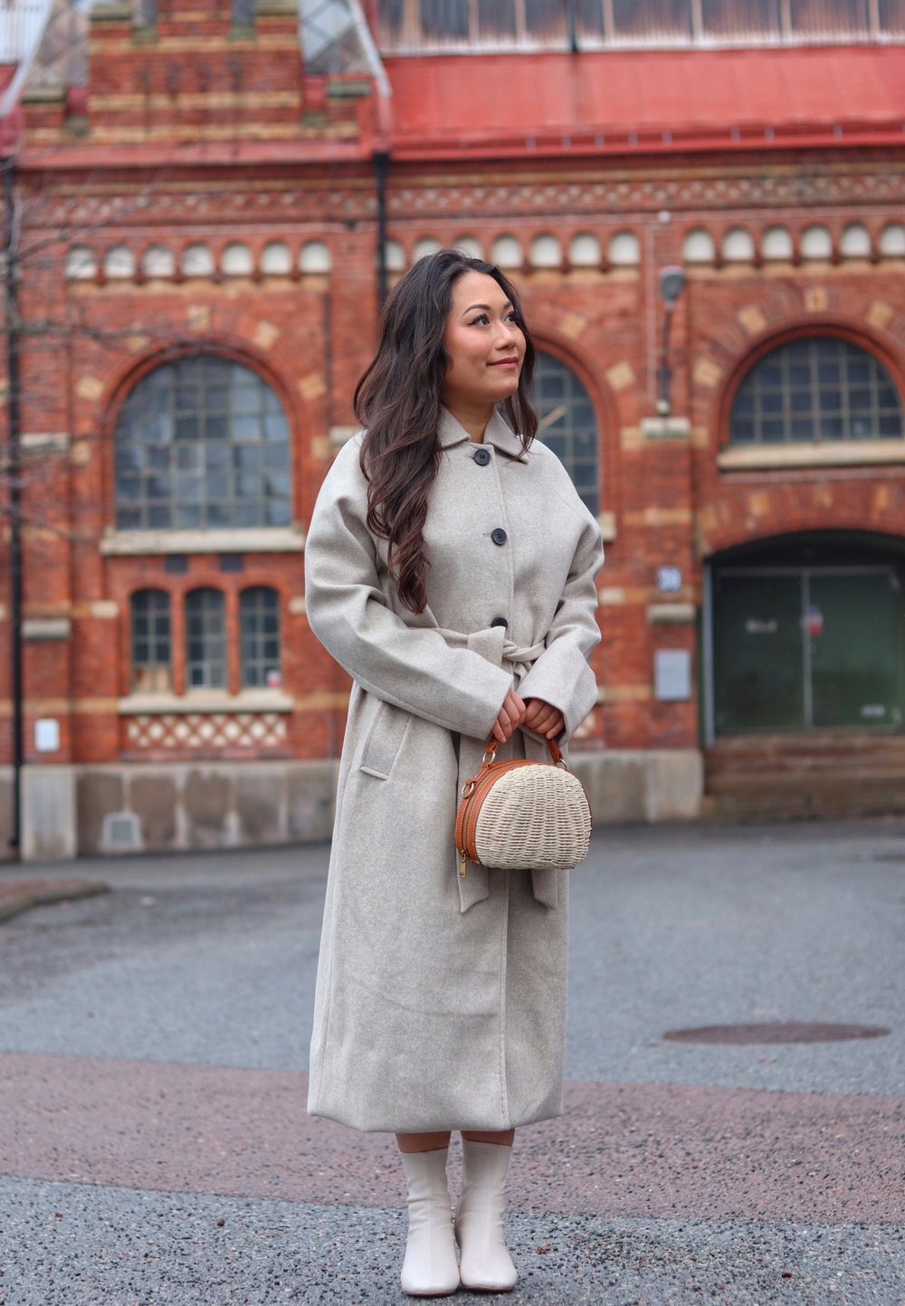 Light gray wool coat with a relaxed fit and belt, complemented by a round wicker handbag with a brown leather strap and white knee-high boots.