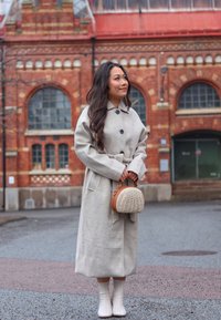 Light gray wool coat with a relaxed fit and belt, complemented by a round wicker handbag with a brown leather strap and white knee-high boots.