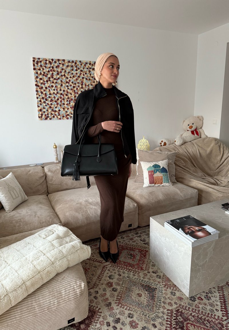 Femme portant un foulard beige, une veste noire et une robe marron, tenant un sac à main noir, debout dans un salon avec des meubles beiges et des coussins décoratifs.
