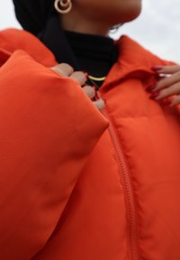 Doudoune orange vif avec un col haut, fermeture éclair à l'avant et texture lisse. Présente une personne ajustant le col.