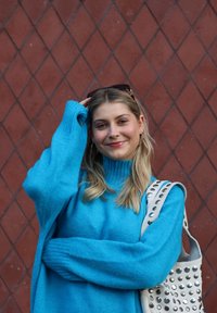 Jasnoniebieski, oversize sweter z prążkowanymi mankietami, zestawiony z kremową torebką ozdobioną okrągłymi metalowymi ćwiekami. Tło ma wzór diamentów.