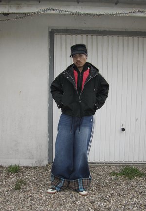Hombre con vaqueros azules grandes con puños de cuadros, chaqueta negra sobre sudadera roja con capucha, gorra negra y gafas, de pie sobre grava frente a una puerta blanca.