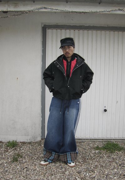Hombre con vaqueros azules grandes con puños de cuadros, chaqueta negra sobre sudadera roja con capucha, gorra negra y gafas, de pie sobre grava frente a una puerta blanca.