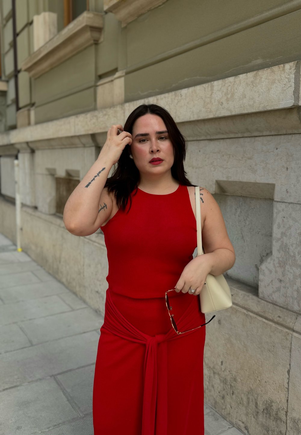 Robe rouge sans manches, ajustée avec un détail noué à la taille. Le modèle tient des lunettes de soleil et un petit sac crème. Cadre urbain avec un fond en pierre.