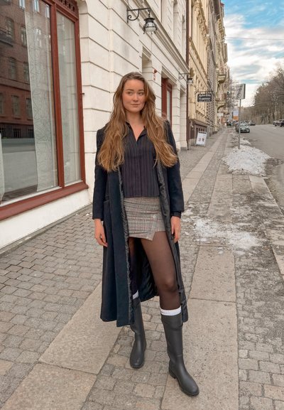 Joven con el pelo largo, que lleva un abrigo negro, camiseta de rayas, falda de cuadros, pantimedias negras y botas, de pie en una acera de la ciudad en invierno.