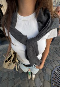 Grauer Pullover um den Hals über einem weißen gerippten T-Shirt gebunden. Helle Jeans und eine beigefarbene geflochtene Handtasche. Trägt mehrfarbige Sneaker.