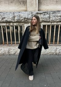 Túlméretezett fekete gyapjúkabát, bézs texturált kötött pulóver fölött, fekete plissézott nadrággal és barna szőrmével bélelt lábbelivel egy köves járdán.
