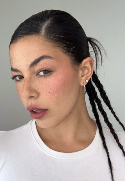 Joven mujer con el pelo oscuro trenzado, pecas claras, lleva una camiseta blanca y varios pendientes, mirando ligeramente hacia un lado sobre un fondo liso.