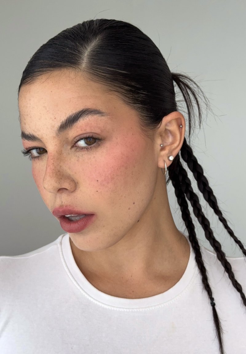 Jeune femme aux cheveux foncés tressés, légères taches de rousseur, portant un haut blanc et plusieurs boucles d'oreilles, regardant légèrement sur le côté sur un fond uni.