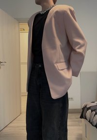 Lyserød blazer med strukturerede skuldre og en enkelt lomme, kombineret med en sort top og mørke denimbukser. Sølvhalskæde synlig.