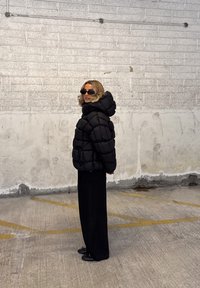 Fekete puffdzseki kapucnival, steppelt mintával. Fekete, bővebb nadrággal és sötét cipővel párosítva, egy texturált fal előtt.