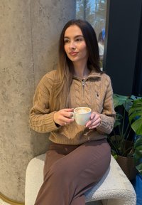 Žena v hnědém svetru a kalhotách sedí na lavičce, drží šálek kávy, poblíž betonového sloupu a zelené pokojové rostliny.