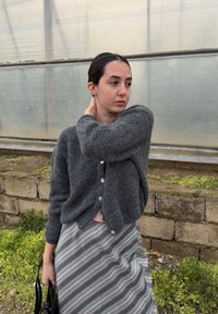 Νεαρή γυναίκα με γκρι ζακέτα και ριγέ φούστα στέκεται έξω κοντά σε πέτρινο τοίχο, αγγίζοντας τον αυχένα της, κοιτάζοντας στο πλάι με ουδέτερη έκφραση.