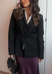 Černé saka na míru s klopovým límcem a zapínáním na knoflíky, doplněné hedvábnou fialovou sukní. Outfit doplňuje malá černá kabelka.