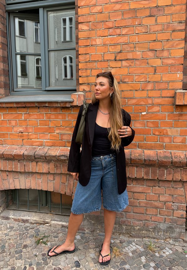 Femme aux longs cheveux portant un blazer noir, un haut noir, un short en jean et des tongs, debout contre un mur de briques rouges près d'une fenêtre.